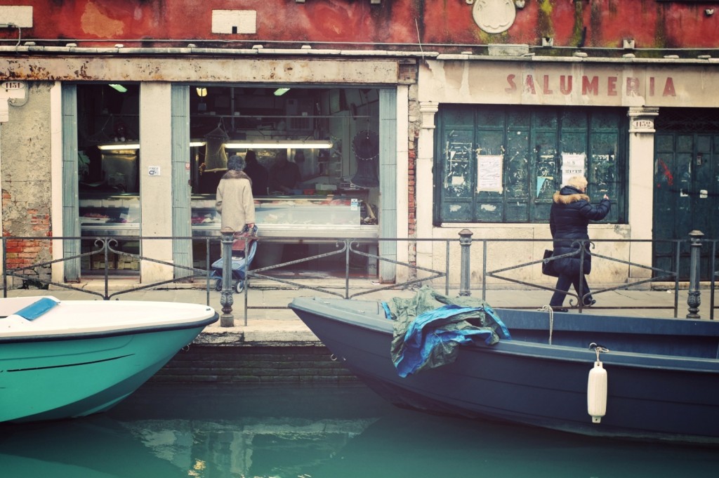 Venezia: quotidianità quieta tra luce fredda, riflessi e presenza umana frontalmente incorniciata dalla vetrina