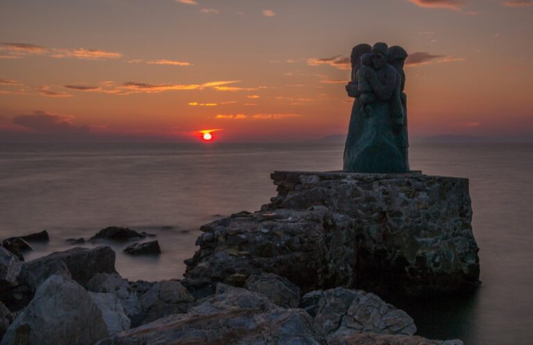 Tramonto che accende la silhouette: mare velluto e scultura sul promontorio