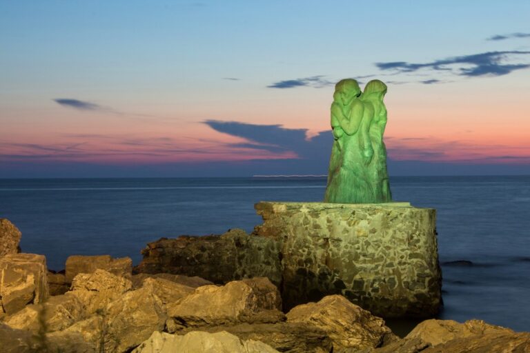 L’Attesa: statua verde e mare al crepuscolo, equilibrio tra roccia e cielo