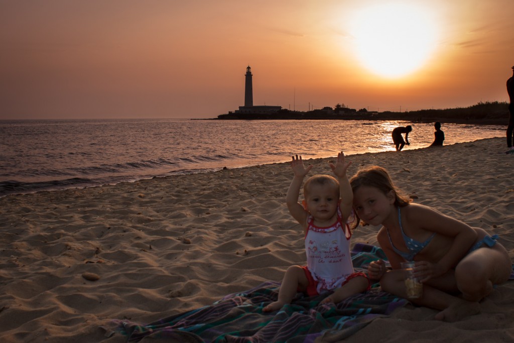 Sguardi in buttero tramonto: gioco tra sorelline, mare e faro