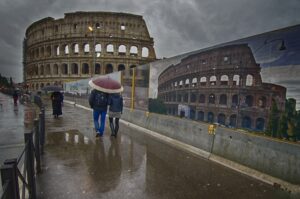 Pioggia su Roma: due passanti tra bara di cantieri e Colosseo in controluce